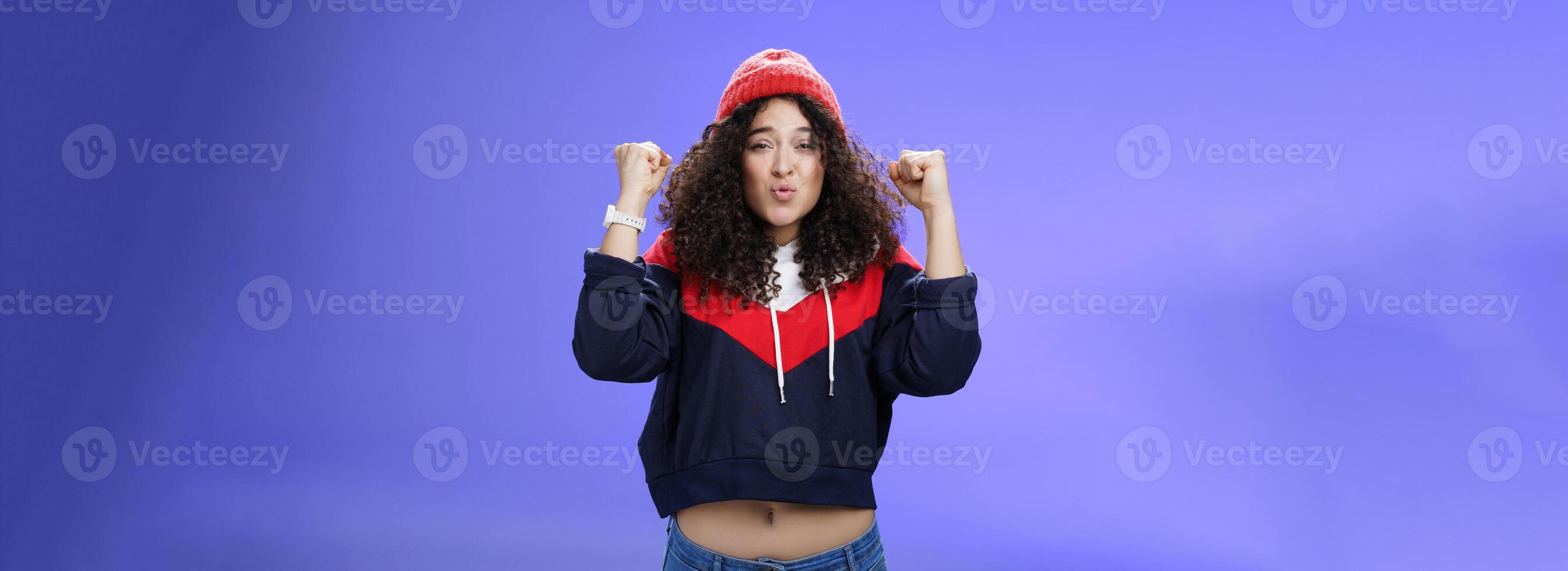 Charming young 20s girl with curly hair in warm beanie raising hands as belly showing under sweatshirt triumphing, celebrating awesome win folding lips from joy and excitement over blue background photo