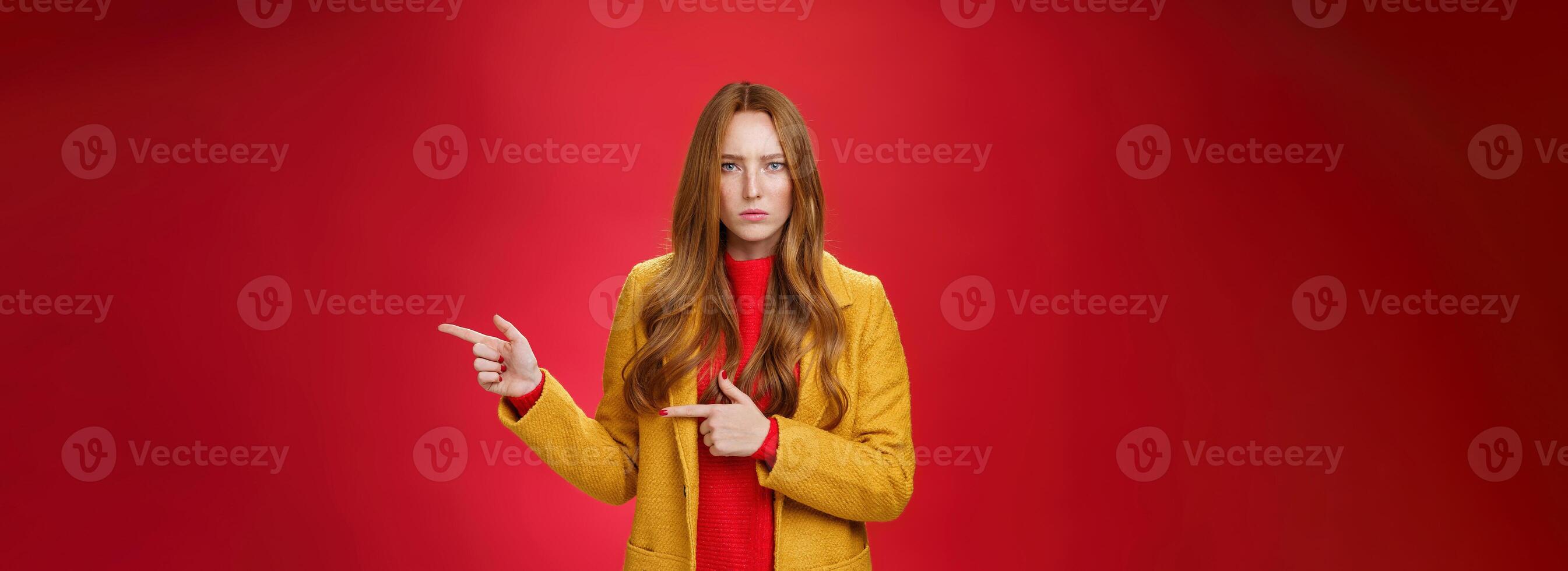 Angry and offended disappointed redhead female in yellow coat frowning looking strict and serious at camera with dislike and agression pointing right asking explain yourself over red background photo