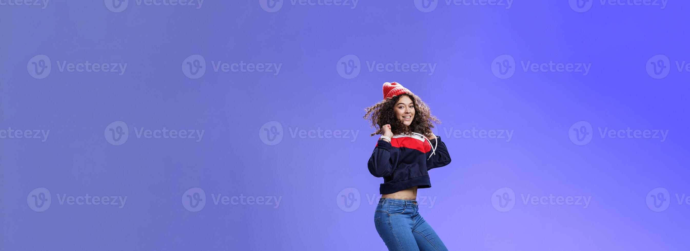 Studio shot of cute girl with curly hair in beanie sweatshirt and sneakers jumping playful and carefree over blue background, having fun enjoying cool weather smiling broadly as flying in air photo