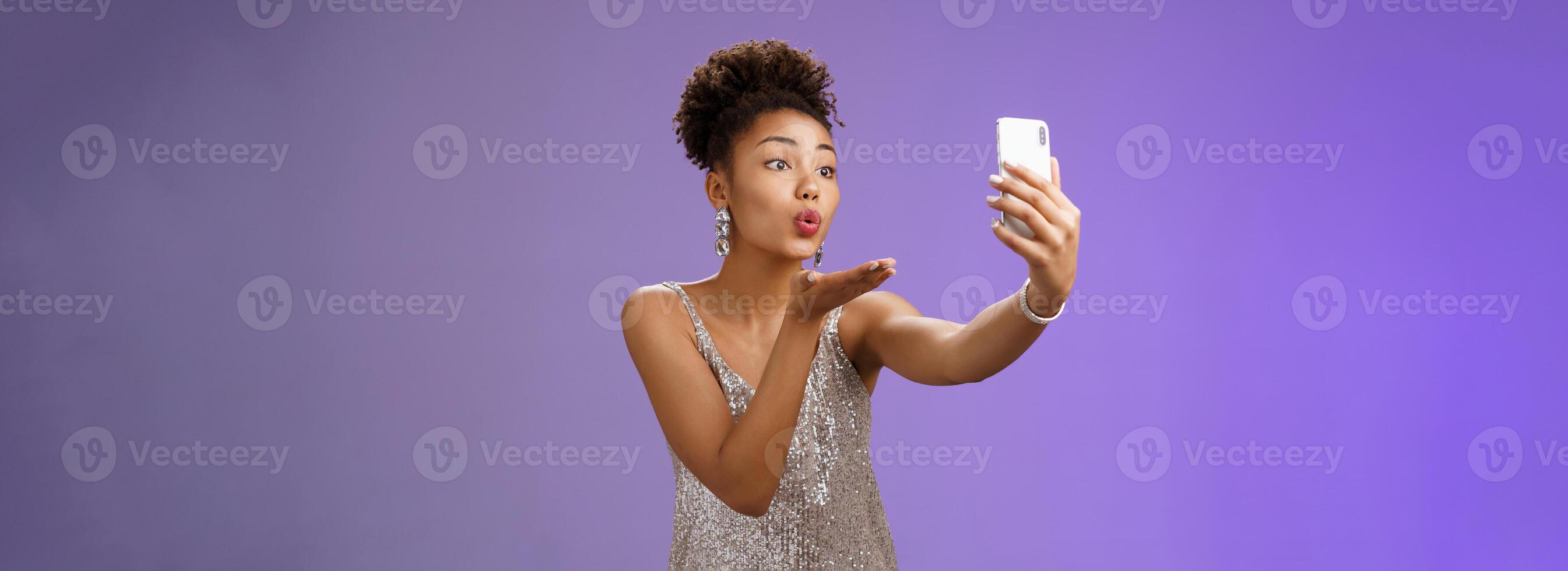Charming elegant young female student partying enjoying prom recording smartphone look phone display send air kiss blowing mwah taking selfie hold gadget, standing blue background photo