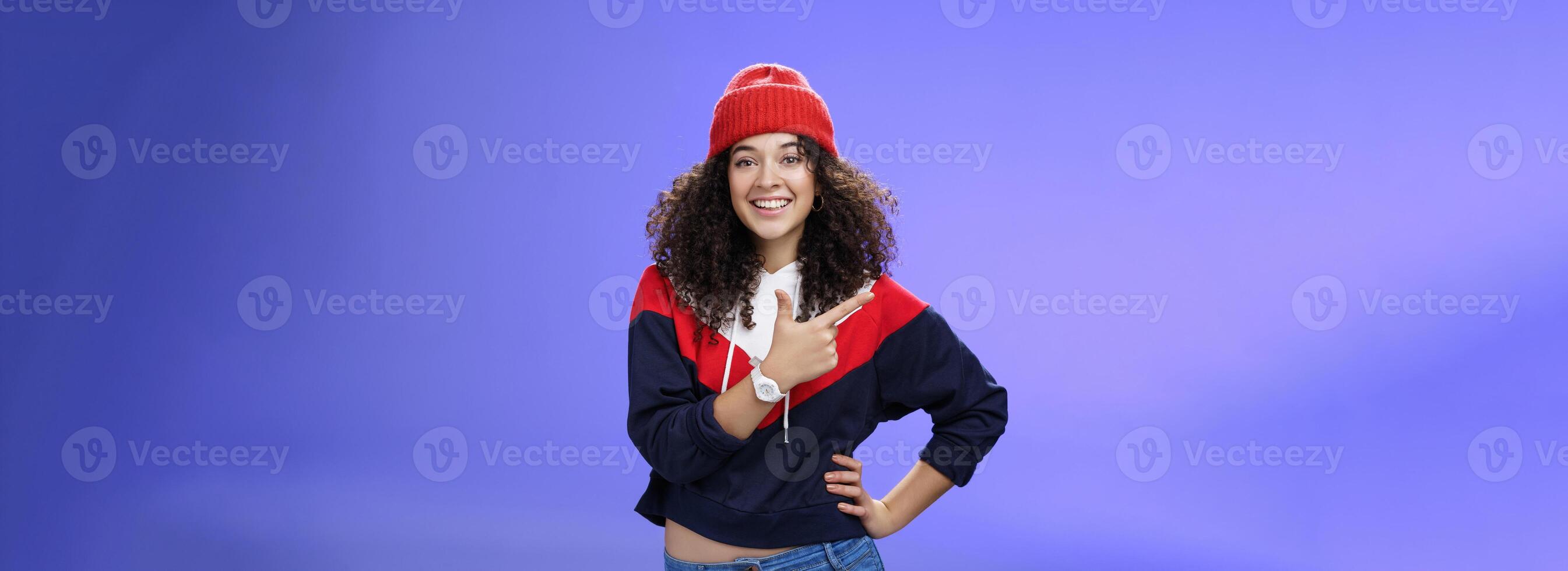 Studio shot of happy charming stylish city girl in beanie and warm sweatshirt ready rock winter up pointing at upper left corner and smiling delighted showing cool place over blue wall photo