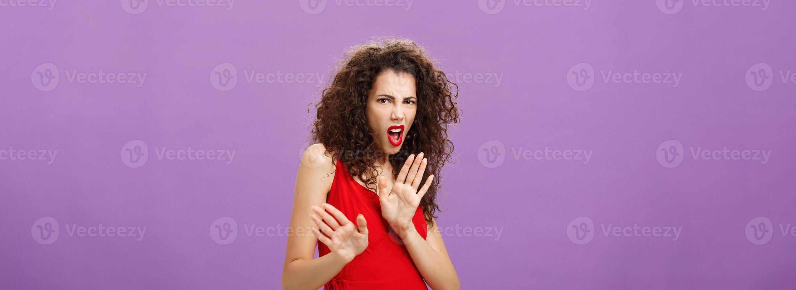 Disgusted arrogant good-looking female with curly hairstyle in red evening dress and makeup pulling palm in aversion and dissatisfaction grimacing rejecting suspicious offer over purple background photo