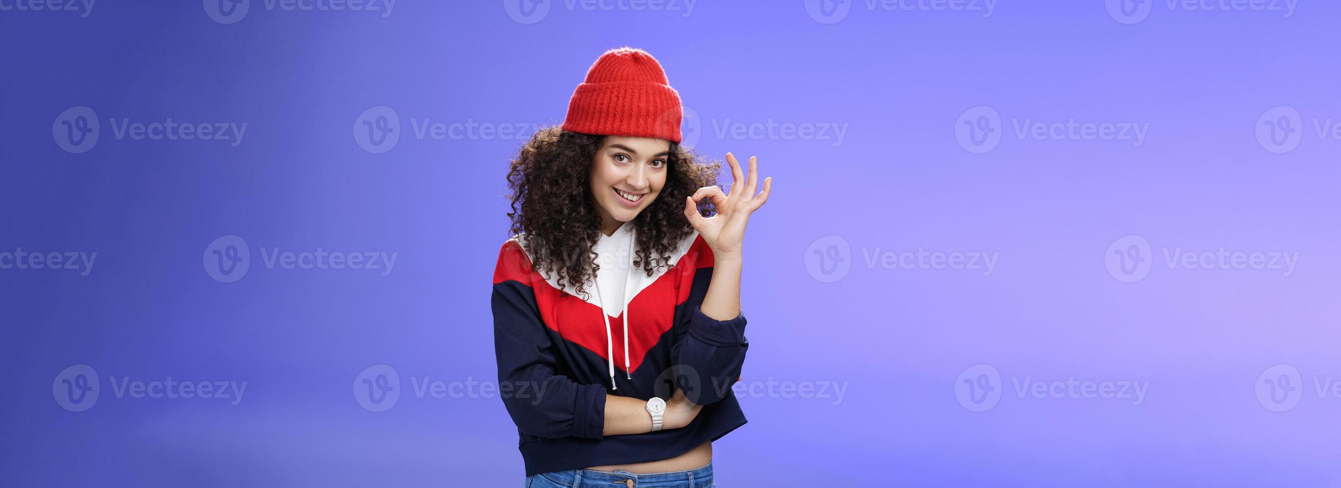 Portrait of pleased good-looking assertive woman with confident look and curly hair wearing warm winter hat showing okay gesture and smiling assuring, liking and approving awesome outfit of friend photo