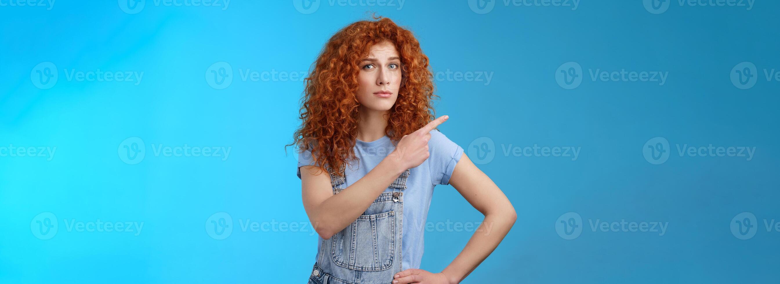 Unsure silly timid hesitant cute redhead curly-haired ginger girl frowning uncertain asking your opinion questioned look camera pointing upper left corner confused worried blue background photo