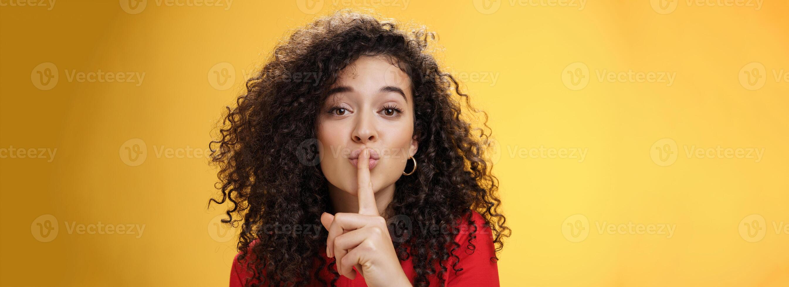 Shh please not tell secret. Cute and tender woman with curly hairstyle gently shushing with index finger on folded lips asking keep promise and remain silent, making surprise over yellow wall photo