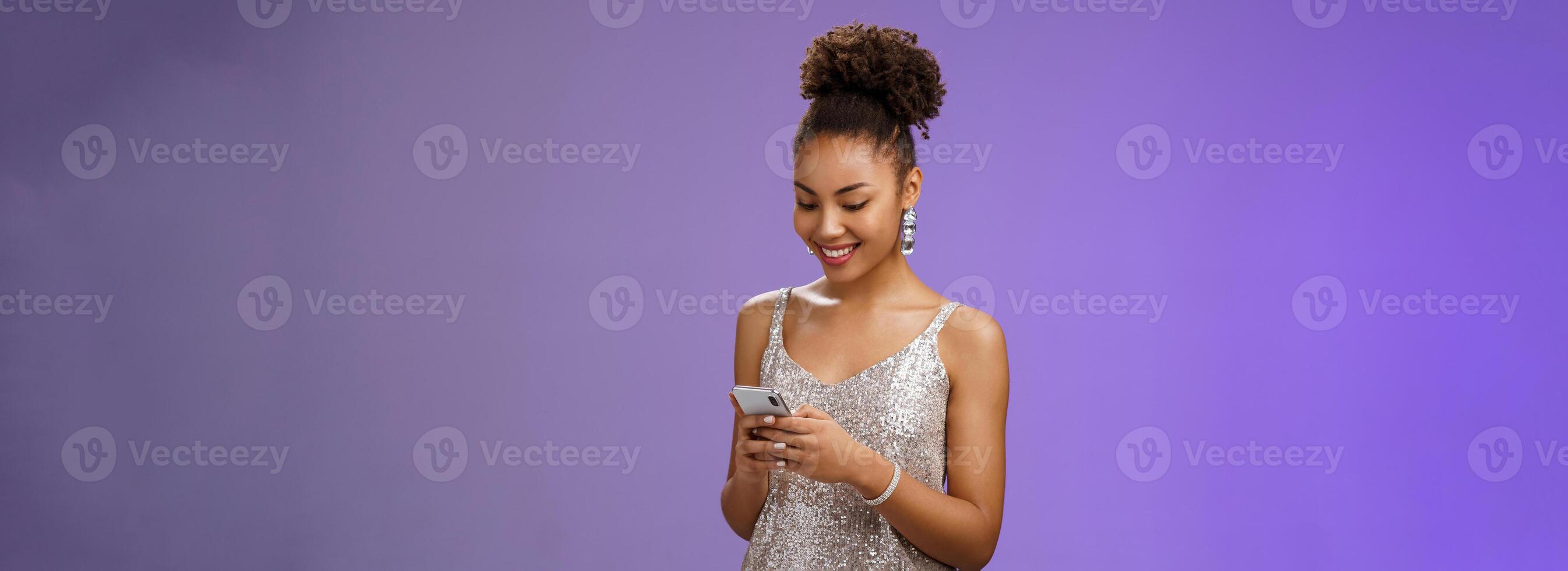 Charming elegant african-american woman in stylish glittering prom dress writing message calling friend hold smartphone look amused happy smile phone display using gadget app scroll party photos