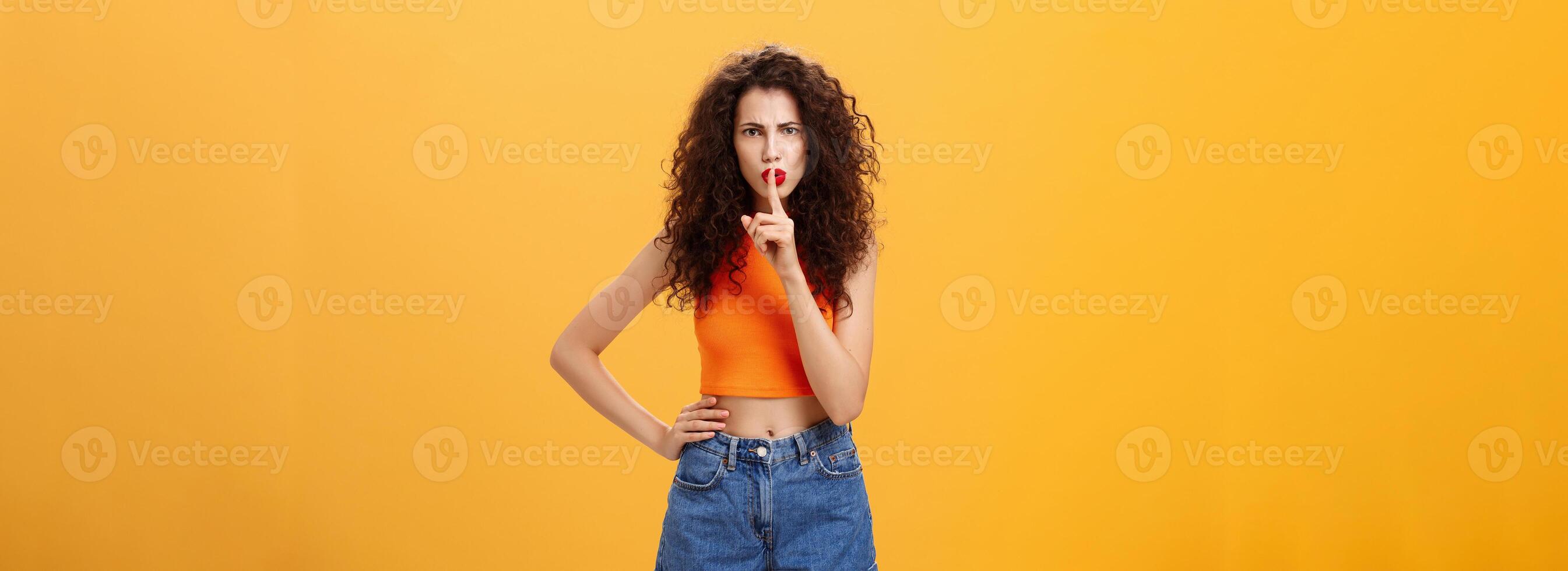 Sister displeased sibling saying cursing words standing serious and strict over orange background in stylish outfit shushing at camera saying shh with index finger over mouth demanding obey rules photo