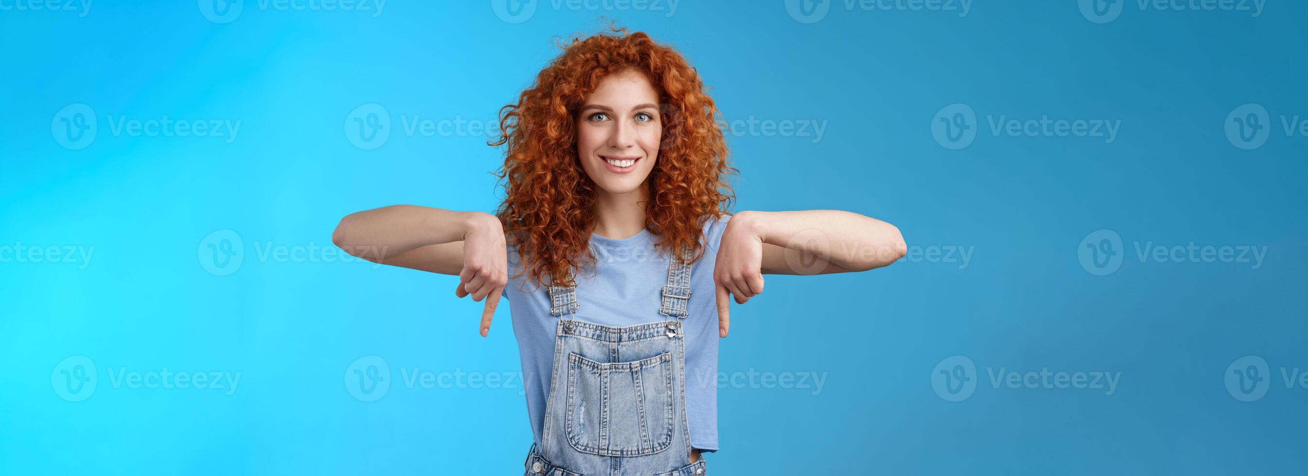 Charismatic sassy flirty redhead daring ginger girl curly haircut pointing down index fingers smiling broadly enthusiastic explore new store pointing promo like cool advertisement blue background photo