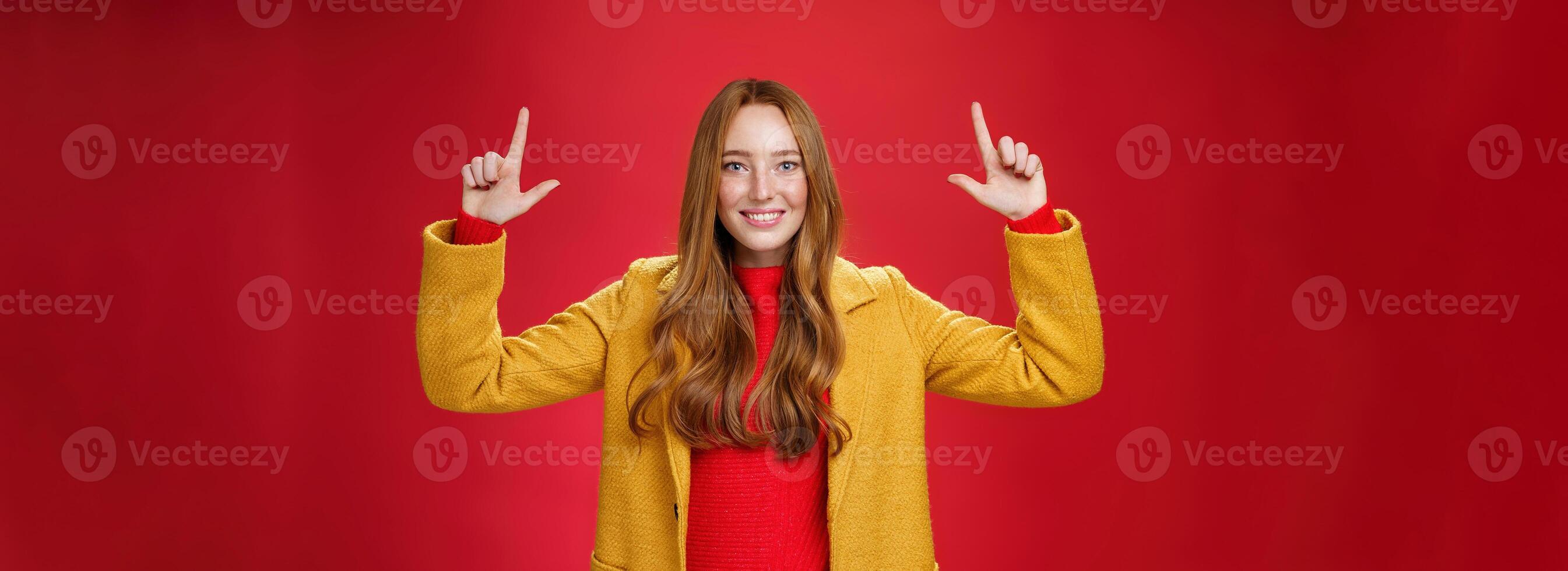 Indoor shot of redhead attractive woman in yellow fall coat raising hands promoting advertisement as pointing up and smiling broadly with satisfied pleasant expression over red background photo