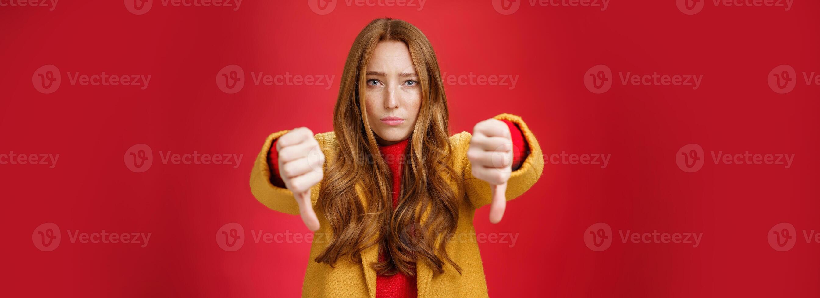 Portrait of insecure and unconfident cute redhead female reacting to uncool thing showing thumbs down with worried upset expression from dislike and disappointment over red background photo