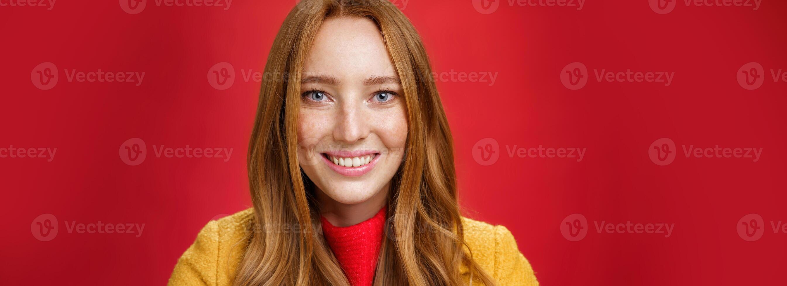 Headshot of hopeful and happy young redhead 20s girlfriend with cute freckles and blue eyes smiling broadly with faithful excited expression having high hopes, posing over red background photo
