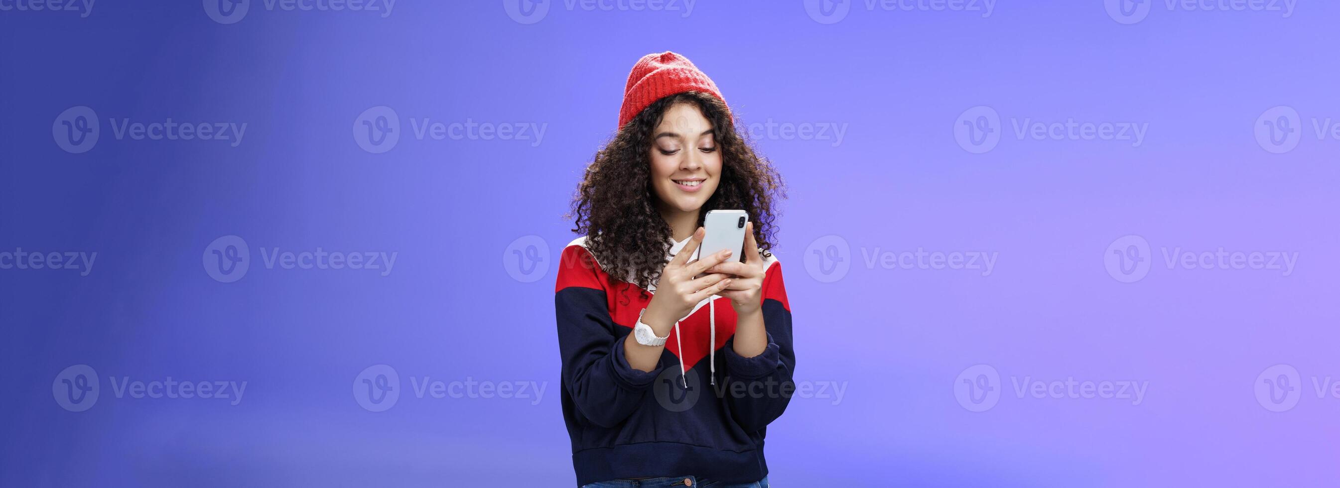 Woman searching in internet funny memes, holding mobile phone in hands smiling joyfully and cute at smartphone screen, typing message or browsing internet over blue background, wearing warm winter hat photo