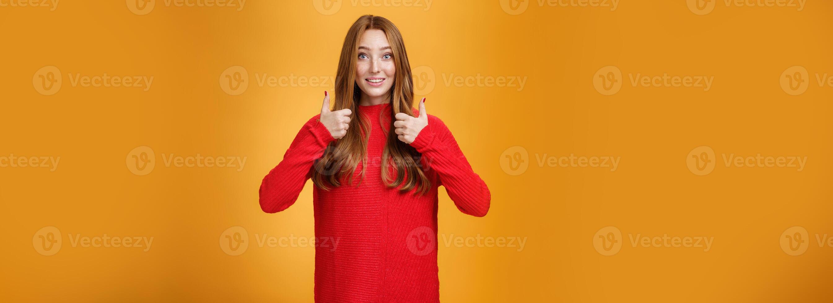 Excited and supportive redhead girlfriend asking opinion about new warm dress showing thumbs up and raising eyebrows questioned as waiting for opinion over orange background photo