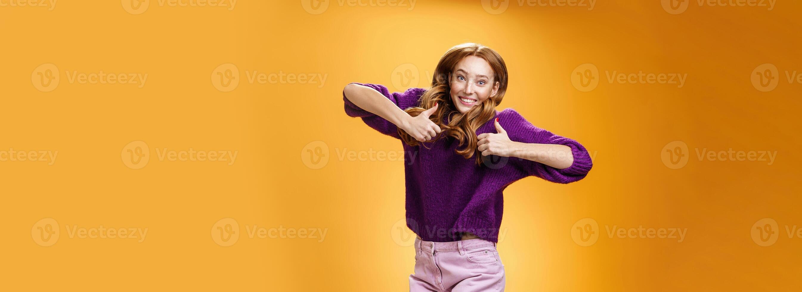 Optimistic and joyful bright ginger girl jumping happily and cheerful having fun showing thumbs up in like and approval giving positive feedback of super cool idea or concept over orange wall photo
