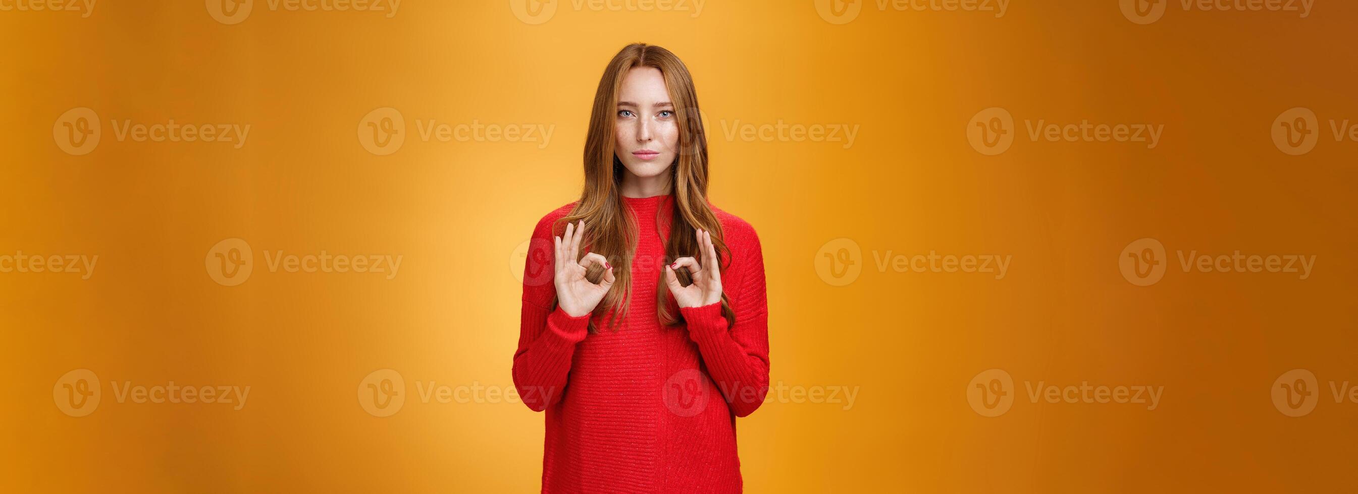 Calm and confident girl showing self-assurance and professionalism as making okay gesture with unemotional expression assuring everything goes by plan standing over orange background, giving approval photo