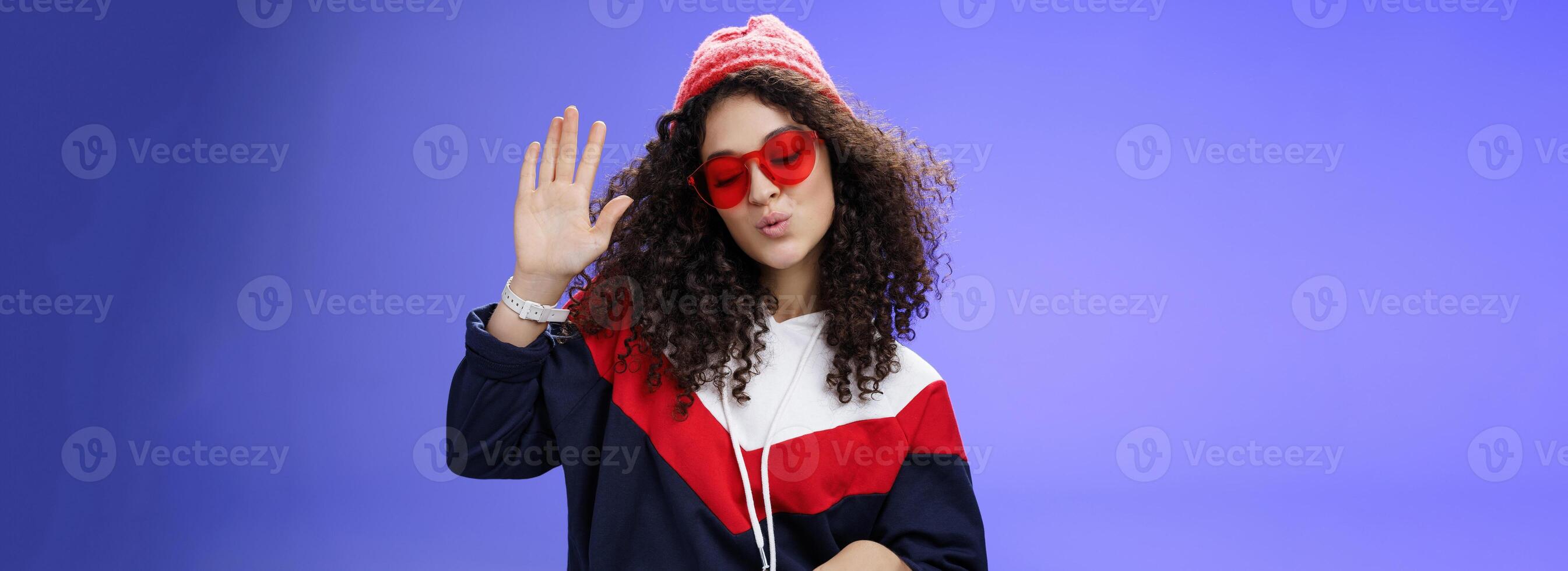 Studio shot of cool and stylish dj girl in red beanie and sunglasses raising palm as enjoying cool music and dancing to rhythm folding lips having fun at awesome party posing chill over blue wall photo
