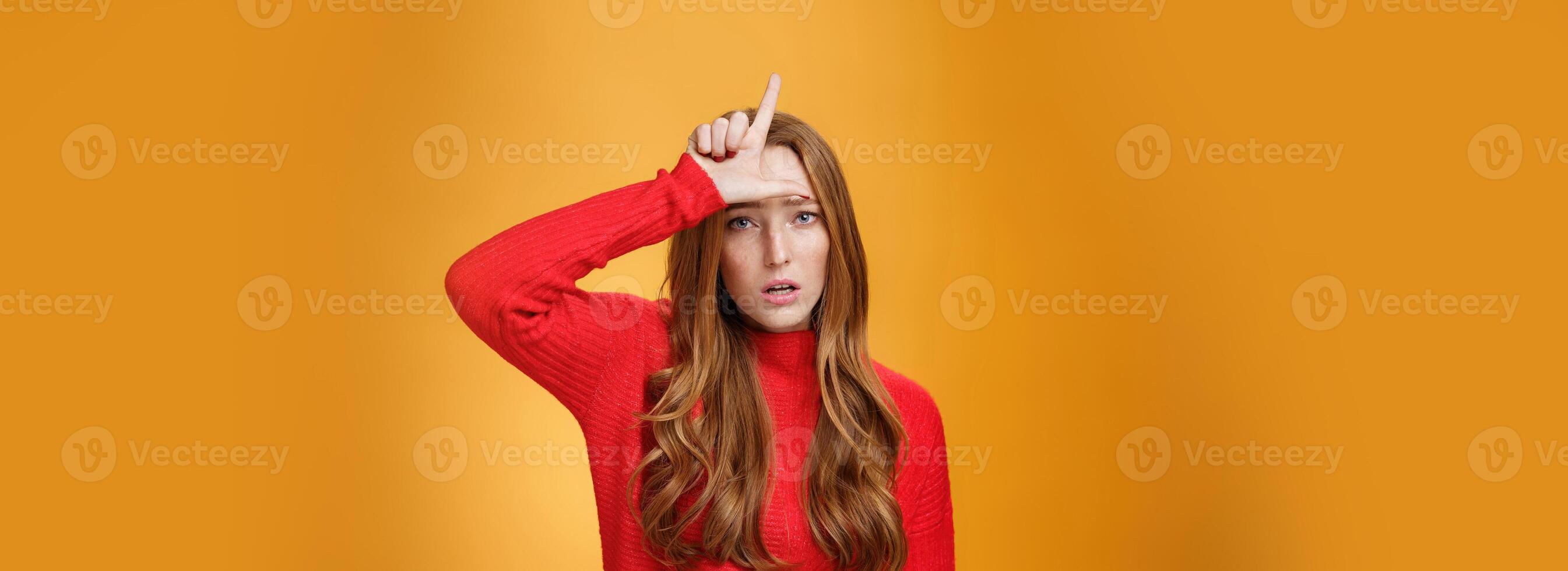 Arrogant and self-satisfied redhead woman humiliating person showing loser sign on forehead mocking and disdain rival standing confident and snobbish over orange background photo
