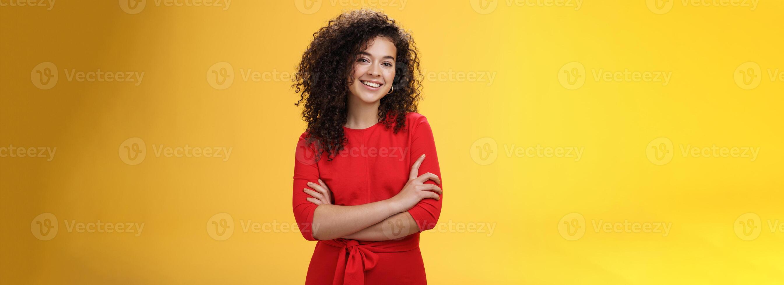 Confident charismatic young curly-haired woman in casual red dress holding hands crossed over chest in self-assured satisfied expression, smiling delighted posing over yellow background photo