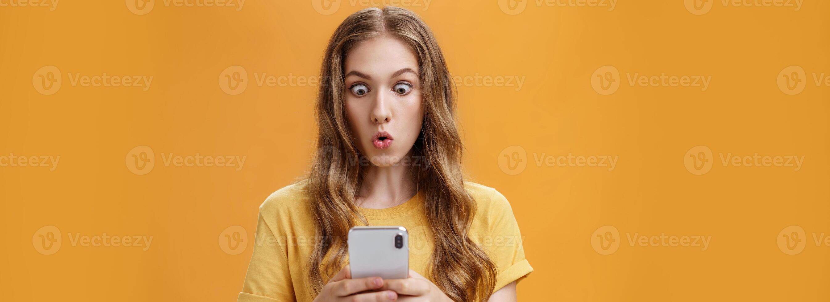 Young girl shocked reading messages in friends smartphone folding lips popping eyes in amazement at cellphone screen folding lips with interest and excitement posing over orange background photo