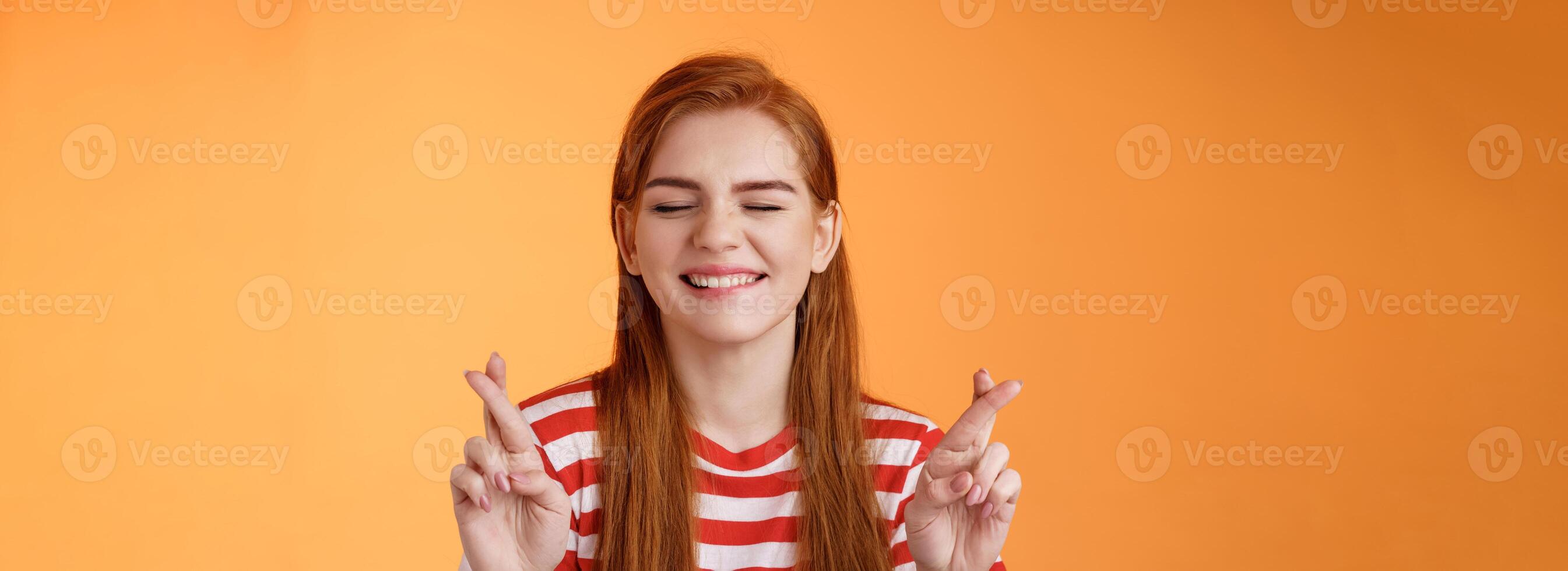 Hopeful optimistic cheerful ginger girl believe dream come true, close eyes cross fingers good luck, making wish, anticipating positive good fortune, stand orange background smiling daydreaming photo