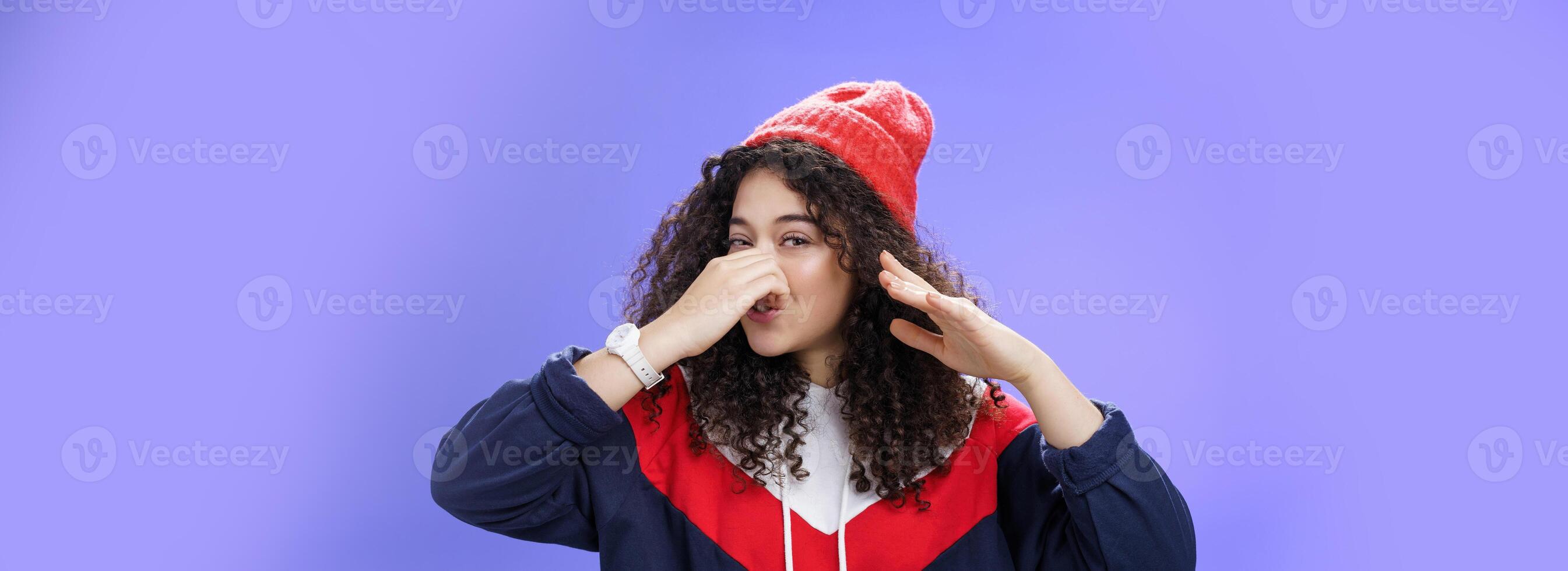 Gosh what stink. Portrait of cute caucasian girl with curly hair in cute trendy hat close nose with fingers and waving hand as smelling awful reek or smell standing disgusted against blue background photo