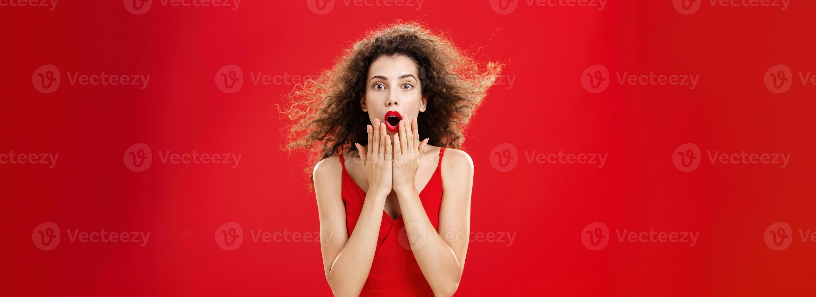 Attractive elegant young woman in red evening dress with curly hair flicking on wind saying wow being impressed and thrilled covering opened mouth from amazement standing over red background photo