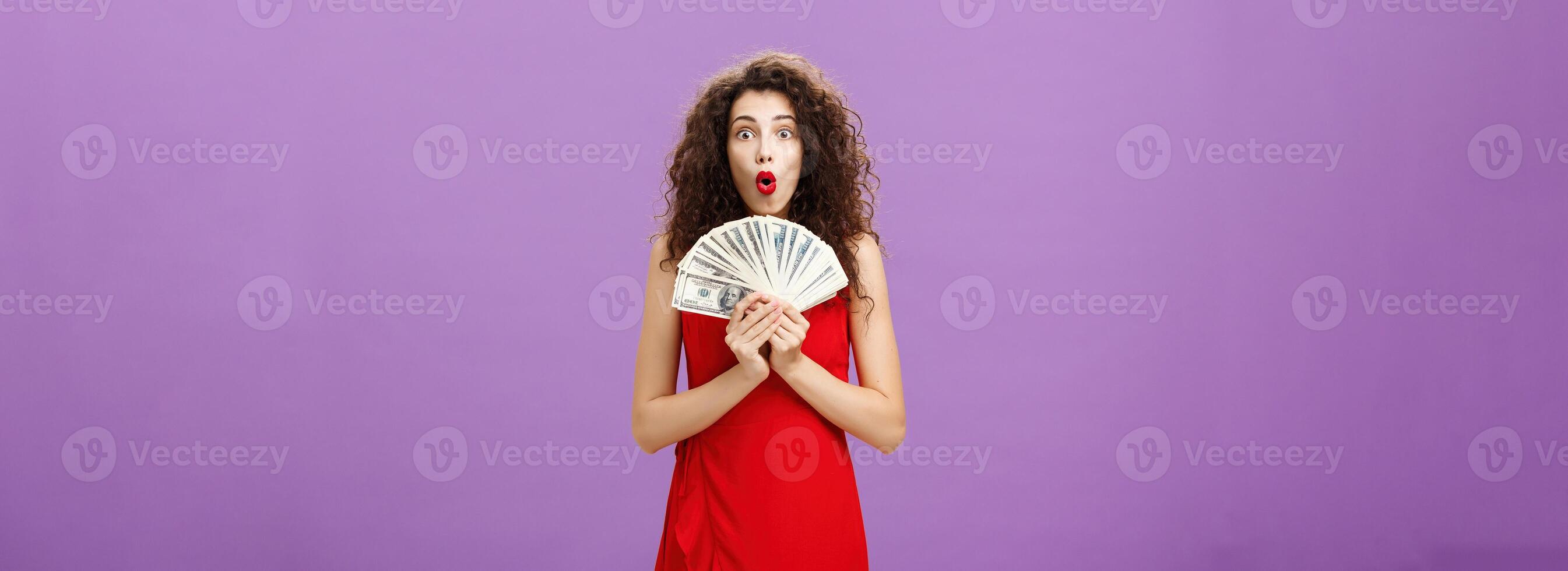 Studio shot of woman being lucky in casino winning lots of cash holding money in hands saying wow with folded lips staring impressed and excited over purple wall in evening red dress photo