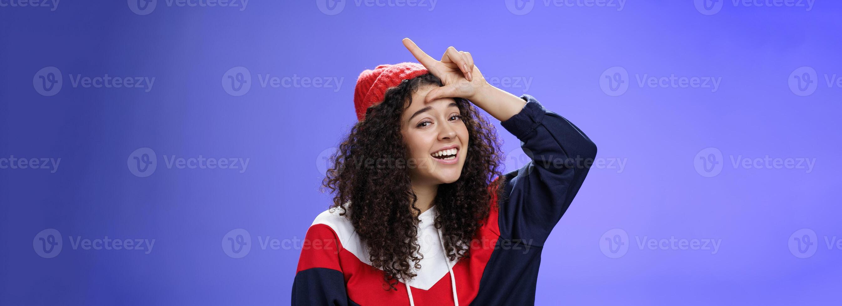 Girl triumphing and mocking friend as winning showing loser gesture on forehead and laughing having fun standing pleased and happy over blue background, smiling at camera joyfully photo