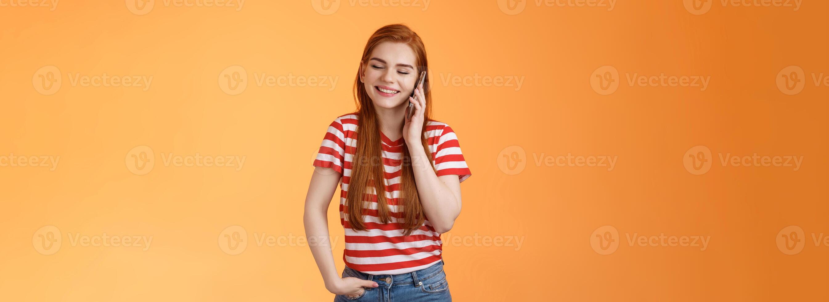 Friendly talkative good-looking redhead female chatting lively discuss ordinary things hold smartphone near ear, close eyes smiling joyfuly, communicating, listen voice message, orange background photo