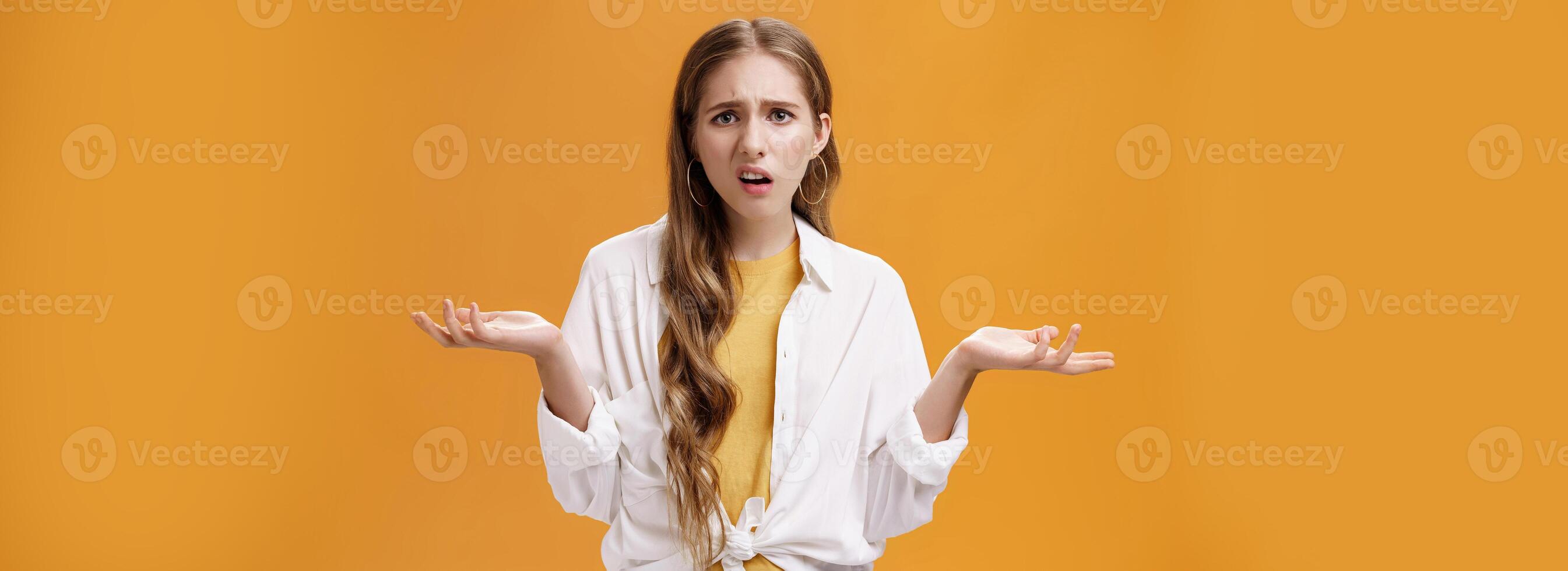 What, I do not understand. Portrait of arrogant questioned impolite girl being pissed with questiones shrugging with hands spread sideways making irritated grimace standing confused over orange wall photo