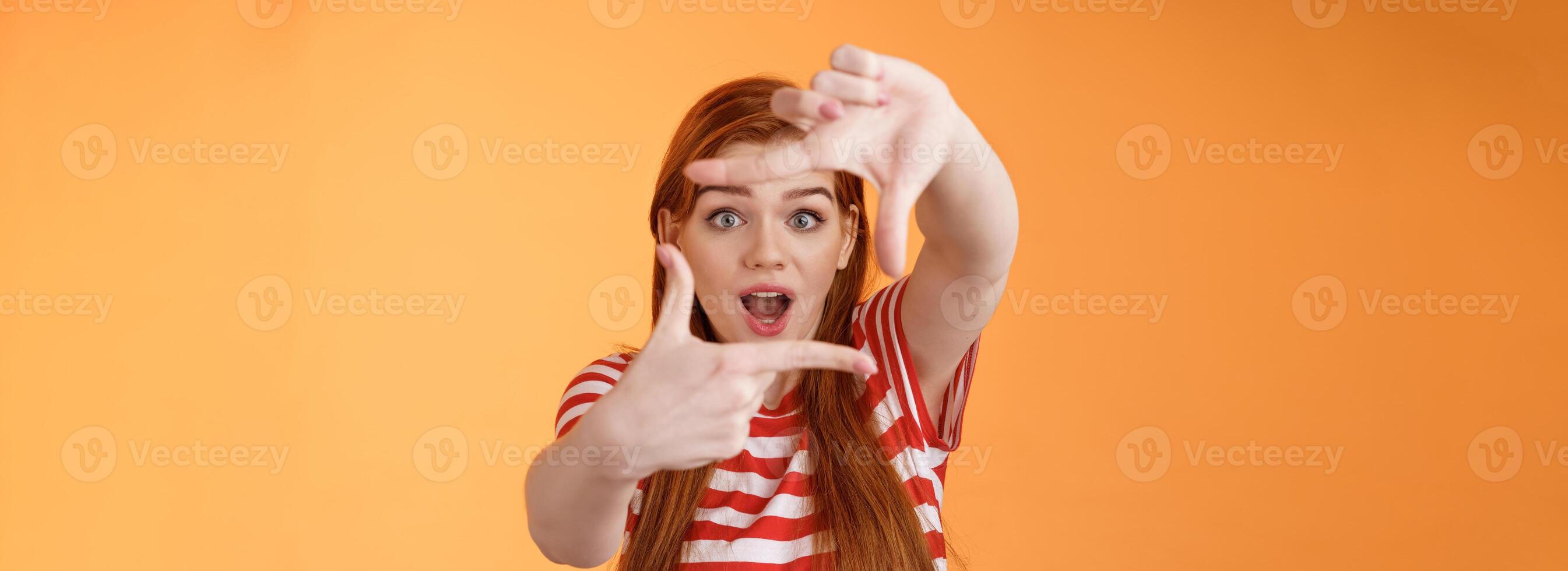Creative upbeat redhead female student got inspiration, imaging how capture interesting shot, extend arms show frames gesture look through hands amazed, open mouth gasping enthusiastic photo