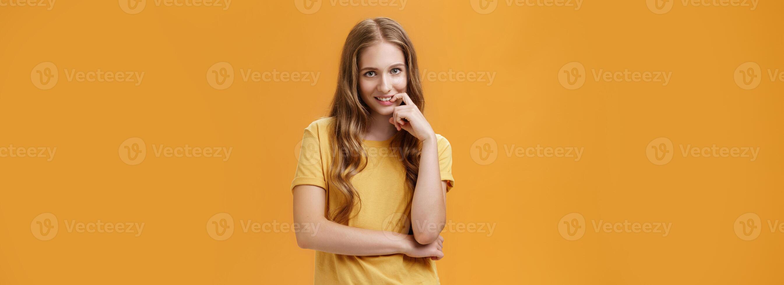 Creative tricky young woman with natural wavy long hair in yellow t-shirt looking from under forehead with intention and lust in expression biting finger, smiling at camera over orange wall photo