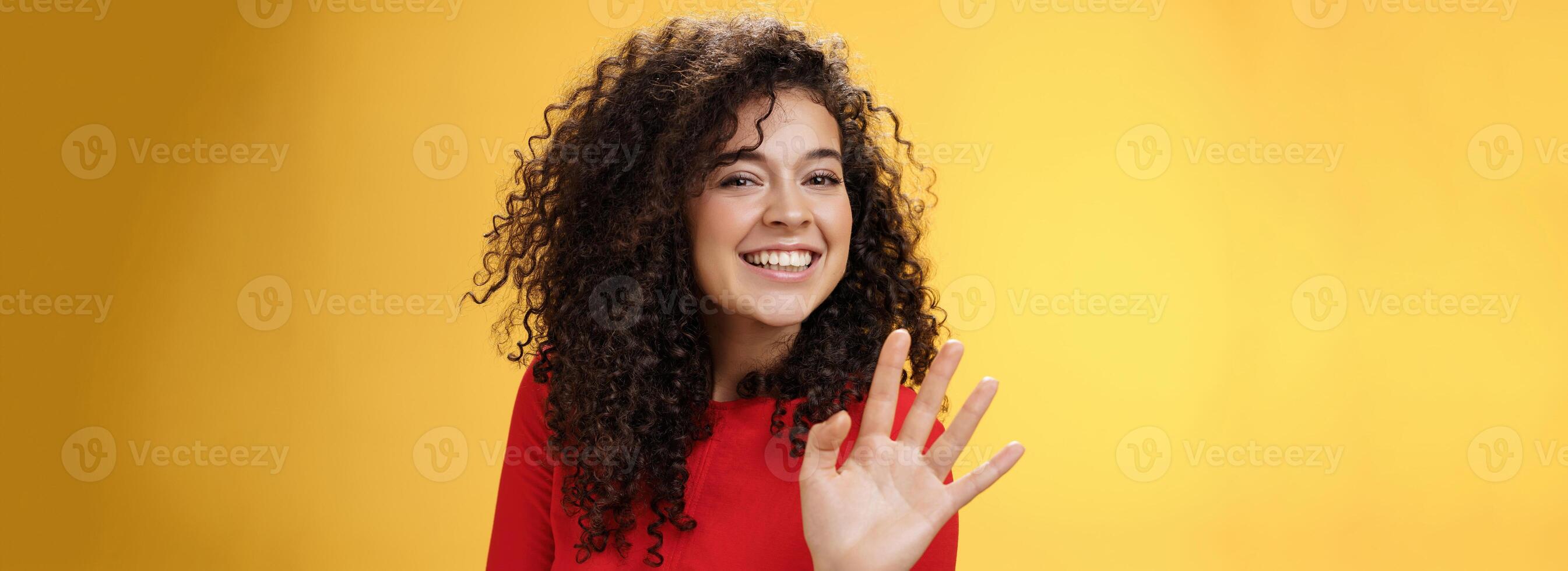 Charming friendly and self-assured attractive curly woman waving cute with palm to say hi or hello smiling broadly greeting man trying flirt in party posing joyful over yellow background photo