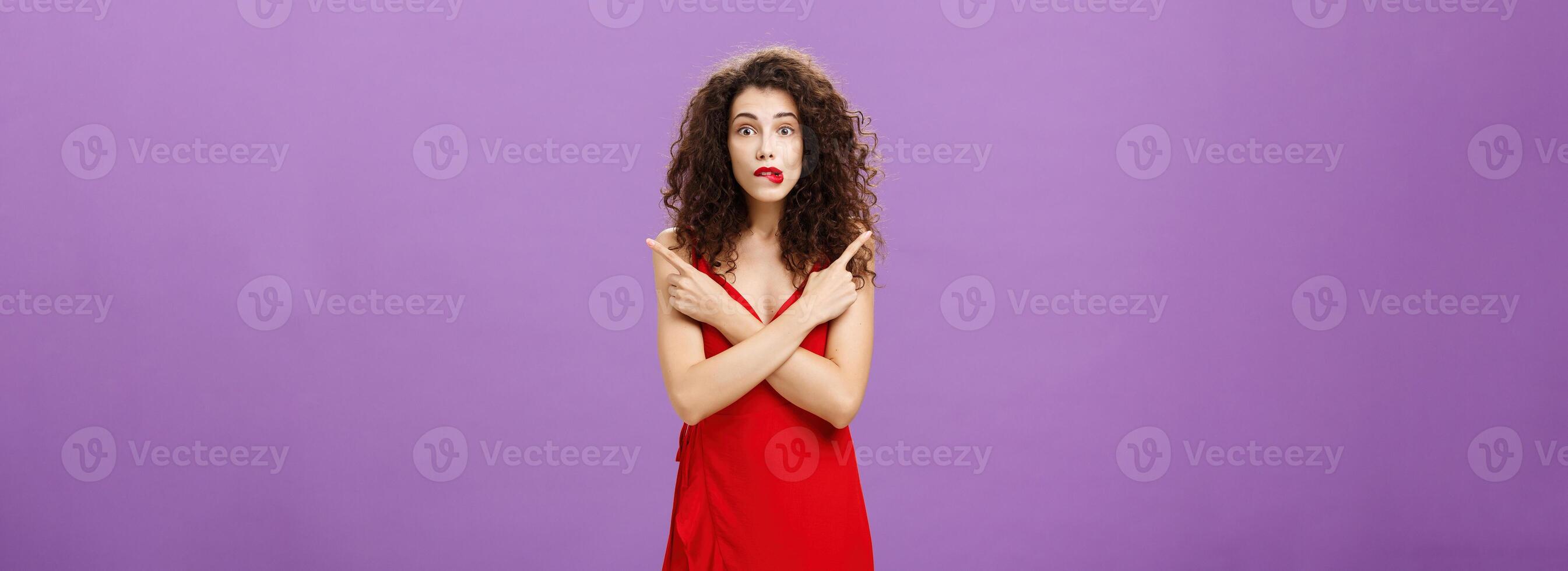 Enthusiastic unsure emotive elegant woman in stylish evening red dress biting lip looking excited pointing in different directions left and right being uncertain and doubtful in making decision photo