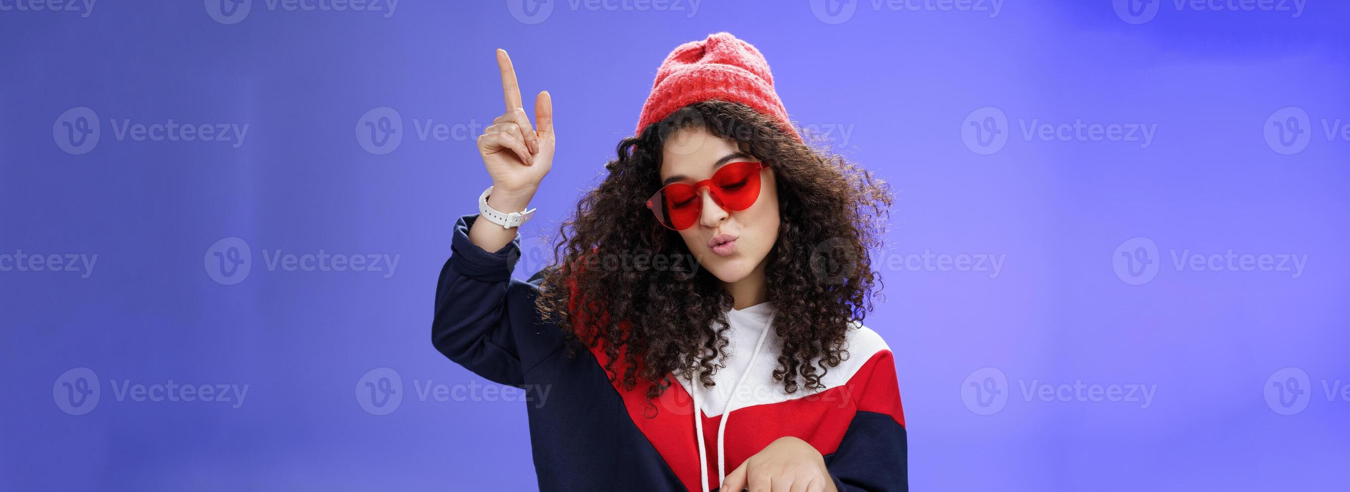 Cute party girl having fun making disco movements folding lips as enjoying cool song dancing joyfully wearing stylish red sunglasses and warm hat looking down enjoying music at party over blue wall photo
