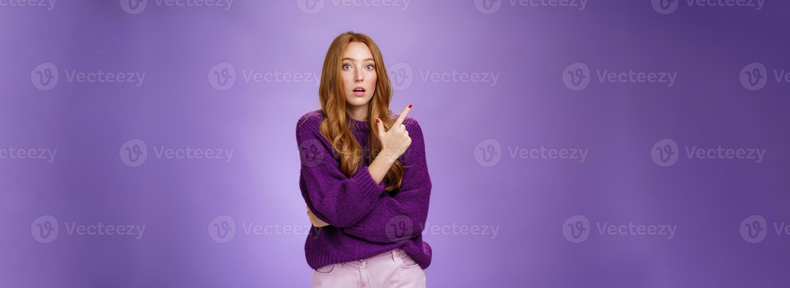 Astonished excited redhead woman open mouth and raise eyebrows questioned as seing shocking thing pointing at upper left corner, asking question impressed and surprised over purple wall photo