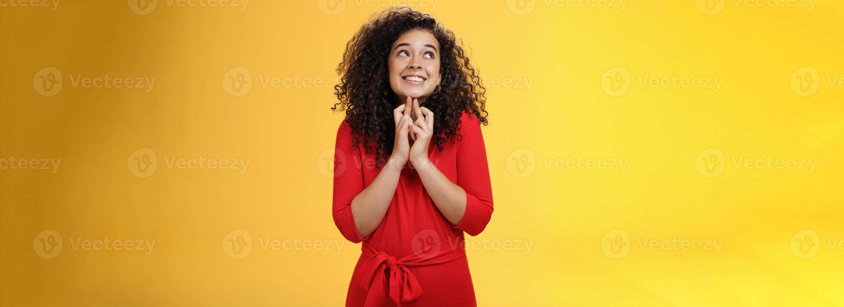 Hopeful, excited cute tender woman in red dress with curly hair standing new x-mass tree crossing fingers for good luck and smiling with head raised to sky making wish, having faith in dream come true photo