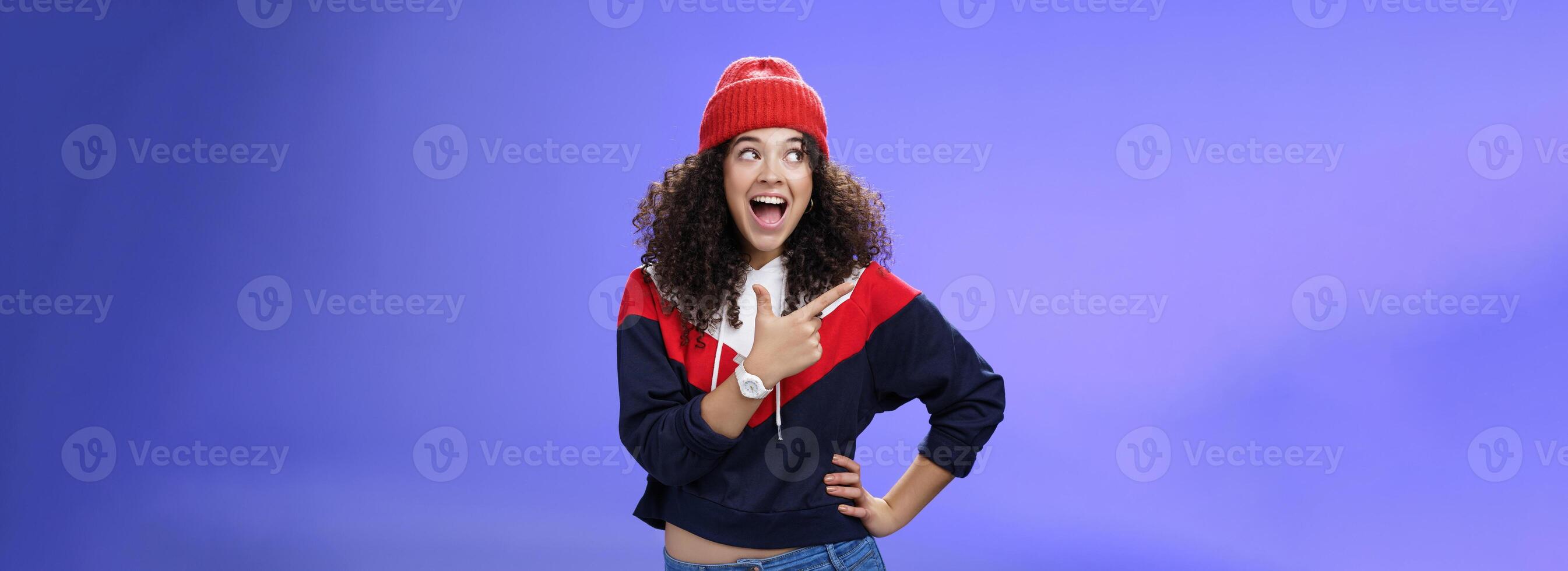 Cute and playful girl in winter stylish outfit and watch smiling broadly with excitement and joy pointing, looking at upper left corner thrilled and joyful as posing over blue background photo