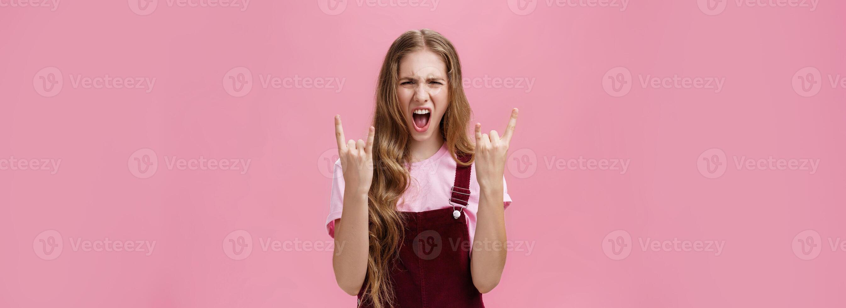 Cute and excited party girl with wavy hairstyle in corduroy overalls yelling yeah and showing rock-n-roll gesture having fun enjoying hard music being daring and rebellious over pink wall photo