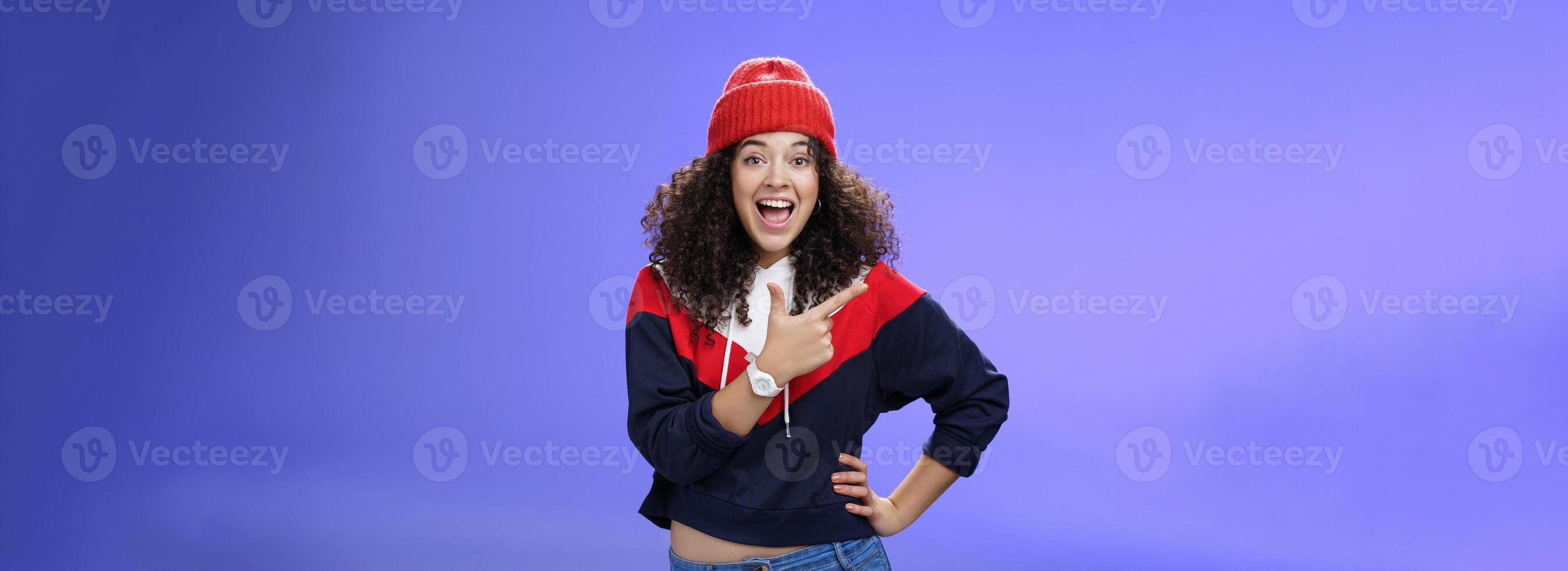 Portrait of enthusiastic and sociable european woman in warm hat and sweatshirt smiling delighted with amused grin as pointing at upper left corner impressed and astonished with awesome promotion photo