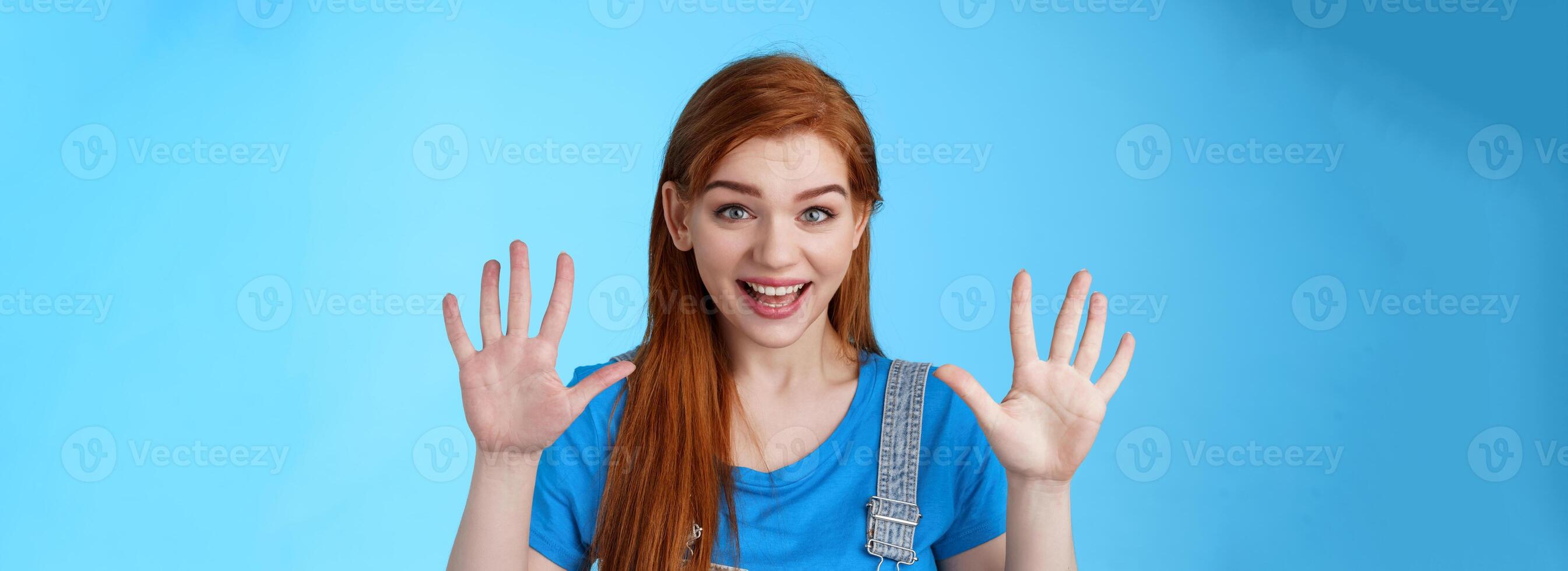 Close-up friendly joyful redhead caucasian girl, long ginger hairstyle, raise hands show number ten, counting dozer, explain fingers, raise arms surrender, smiling joyful, stand blue background photo