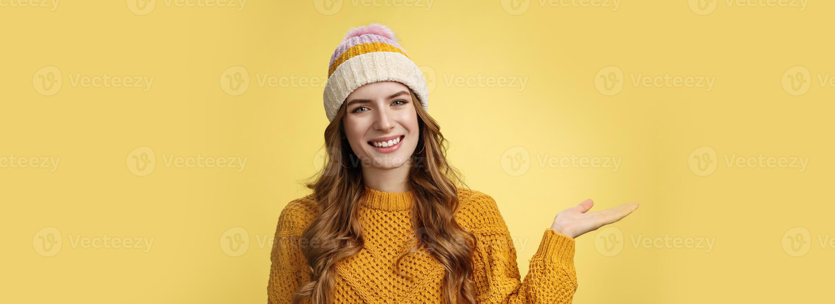 Friendly-looking confident pleasant attactive young european woman wearing winter hat, sweater extend arm show product welcoming inviting guests come in smiling happily, promoting copy space photo