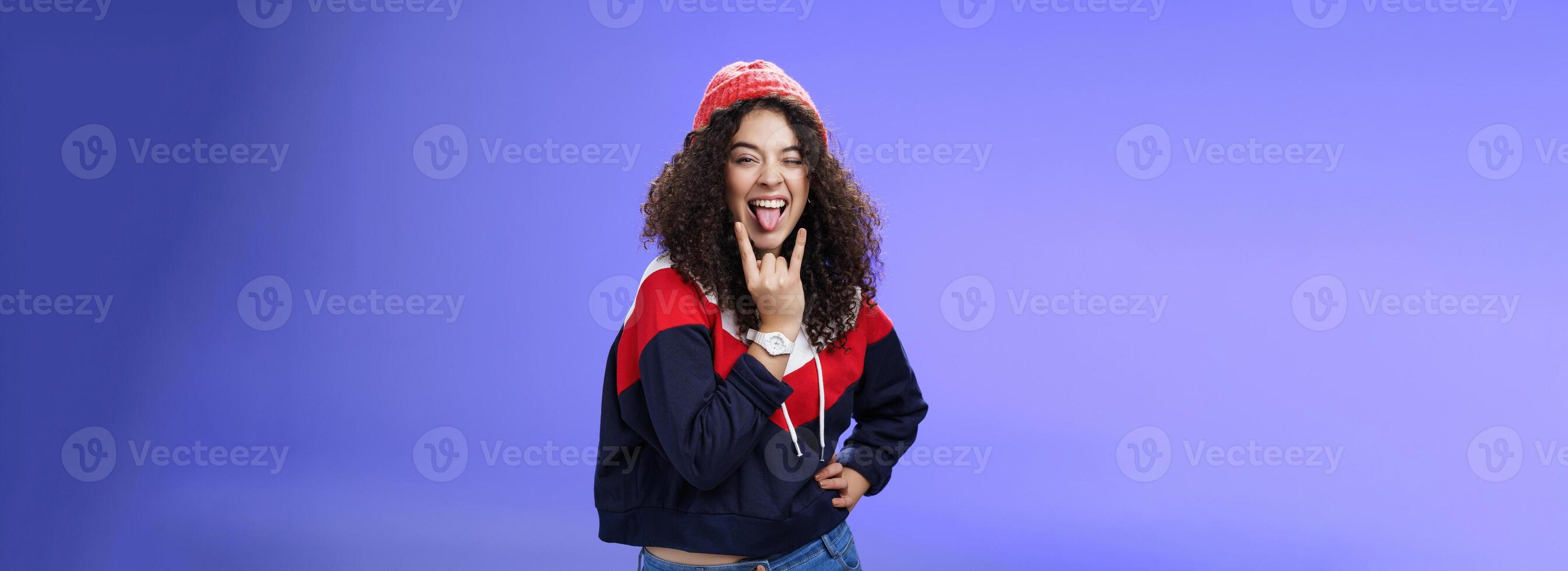 Stylish and cool energized swag girl with curly hairstyle in warm beanie showing rock-n-roll gesture near mouth and sticking out tongue smiling having fun, partying and enjoying life over blue wall photo