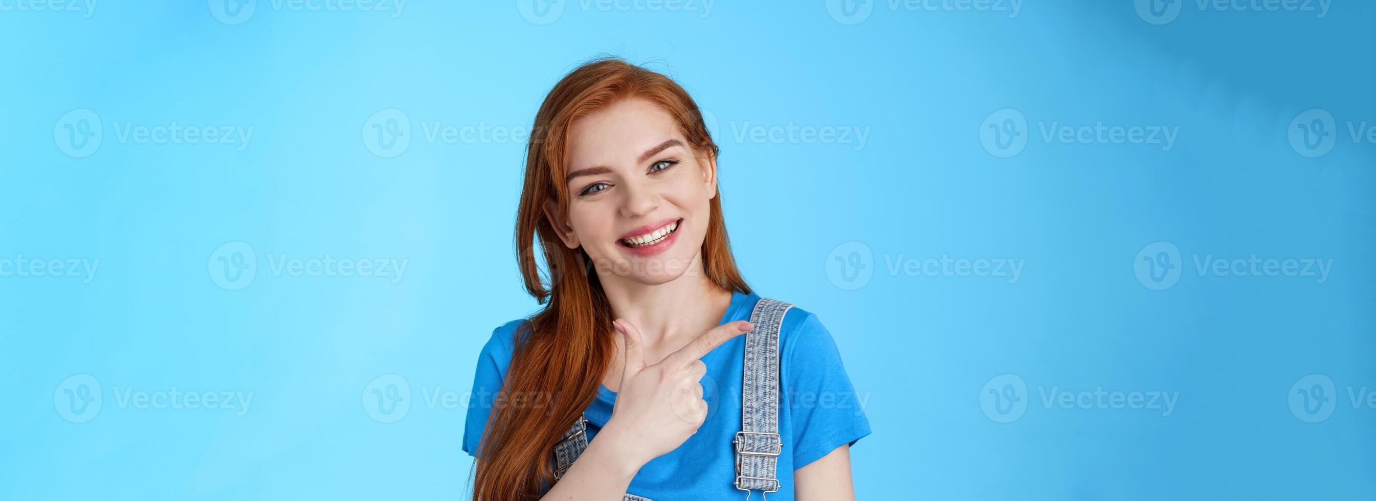 Friendly cheerful caucasian red woman, tilt head smiling pleasantly, suggest good copy space, pointing left introduce promo, give advice, indicating awesome link, pointing blue background photo