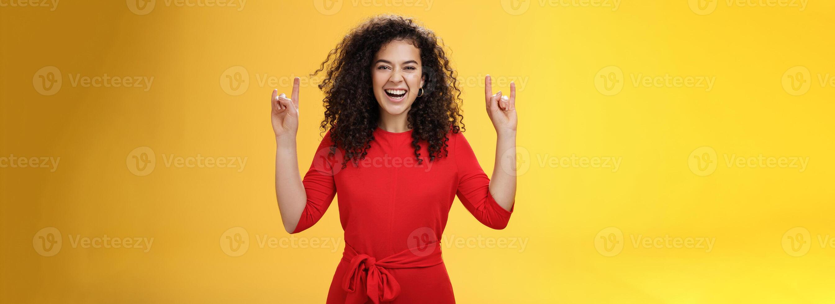 Rock n roll live. Charismatic excited young curly-haired woman in red dress feeling awesome as waiting concert of favorite band showing rock gestures with raised hands and smiling thrilled and happy photo