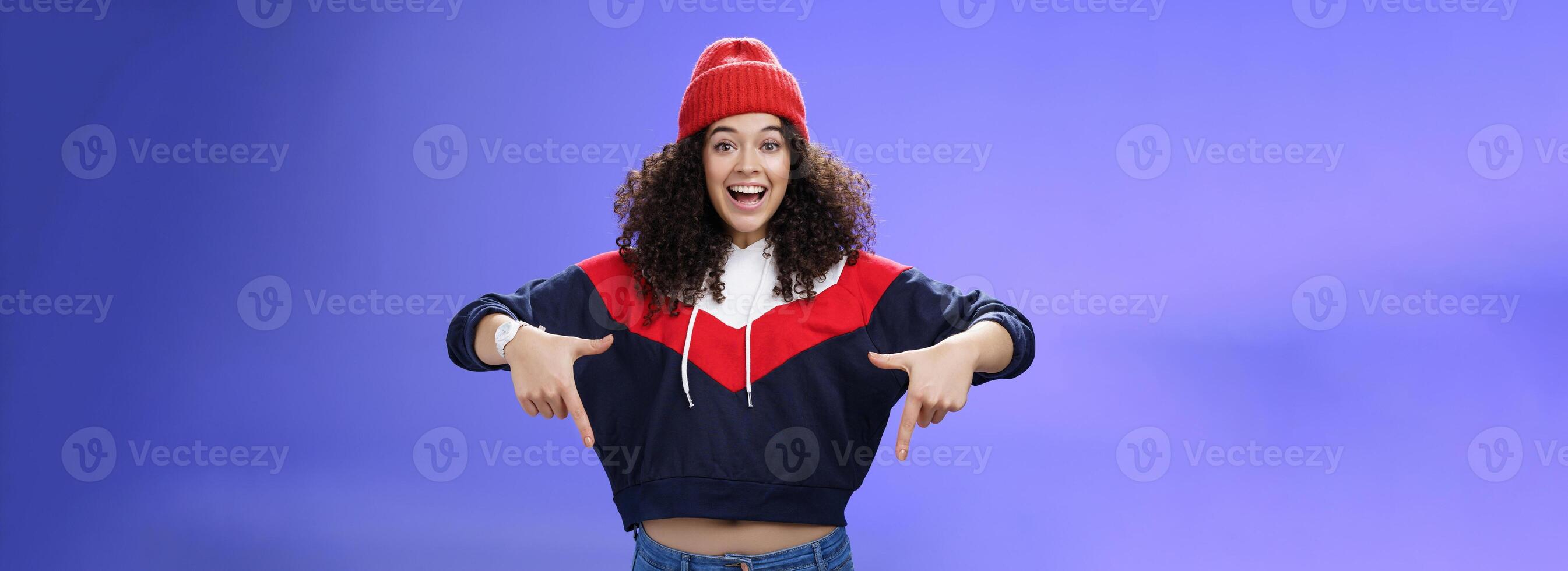 Hurry up and chek it out. Portrait of charismatic joyful and excited young cute curly-haired girl in beanie and stylish outfit pointing down, smiling broadly inviting join fun over blue background photo
