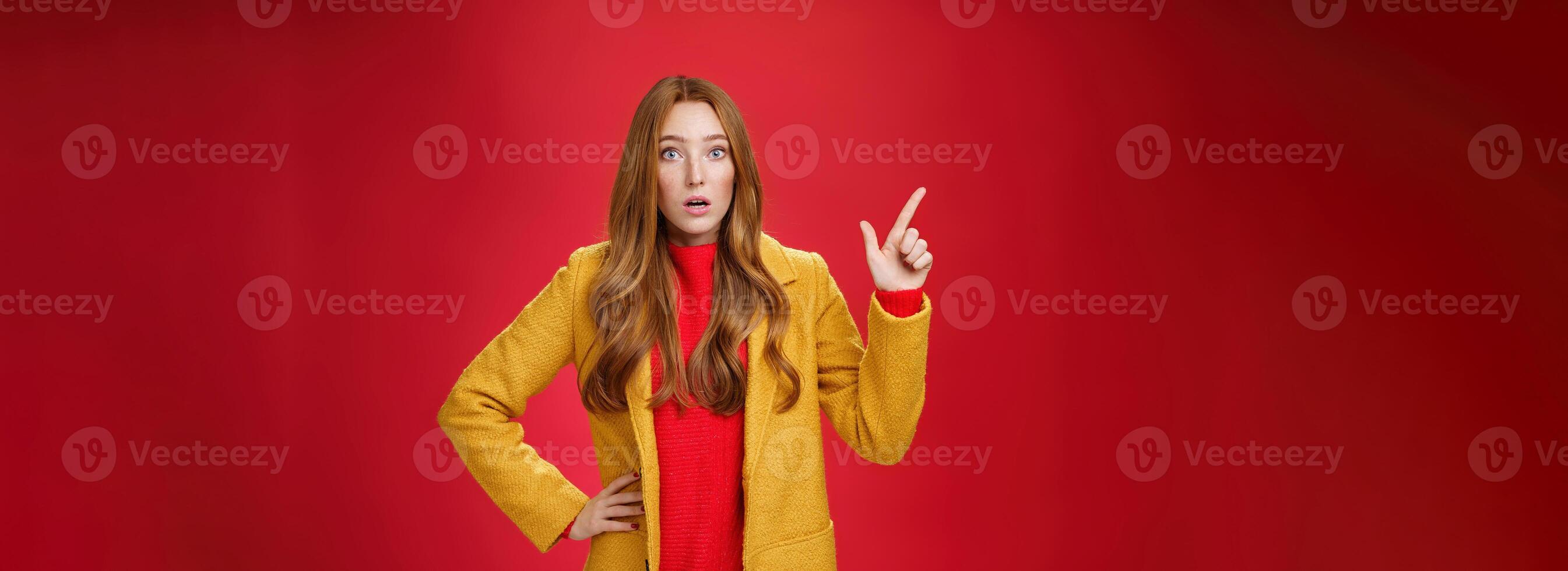 Shocked and stunned cute, confused ginger girl in yellow coat feeling worried and surprised open mouth lifting eyebrows amazed pointing at upper left corner, posing against red background photo