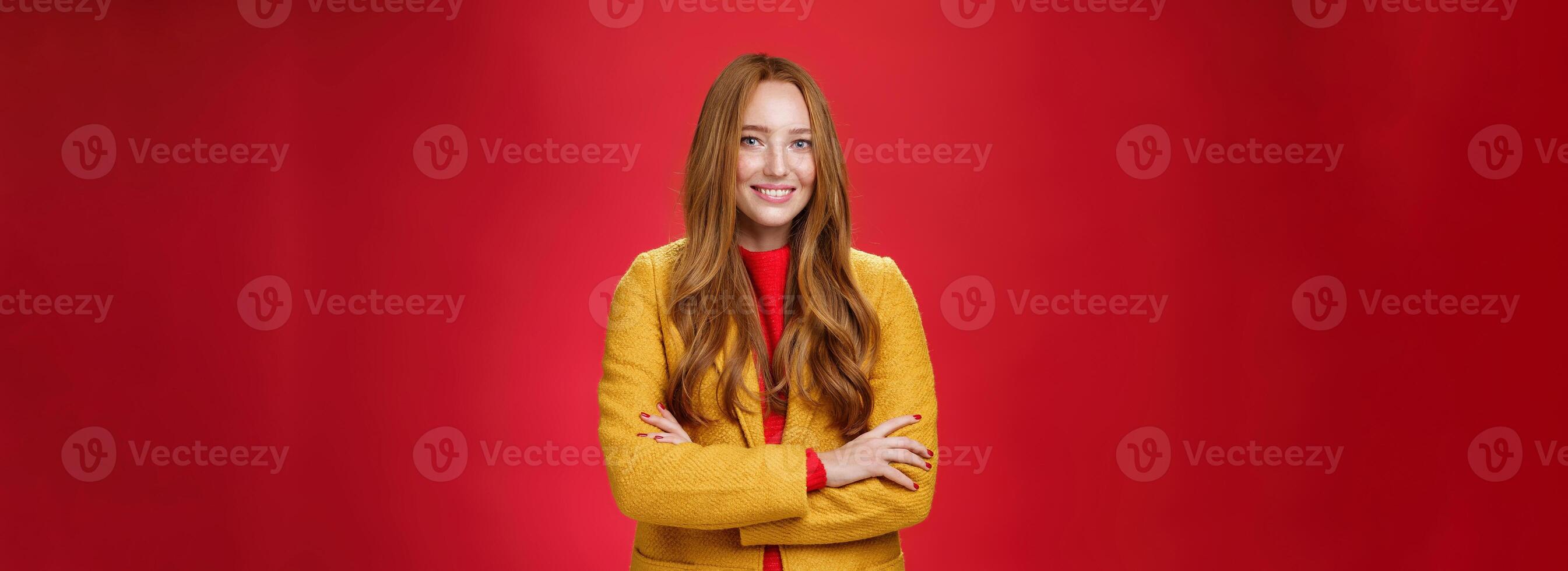 Charming successful young female in yellow autumn coat smiling broadly holding hands crossed on body in confident self-assured pose standing delighted and happy over red background photo