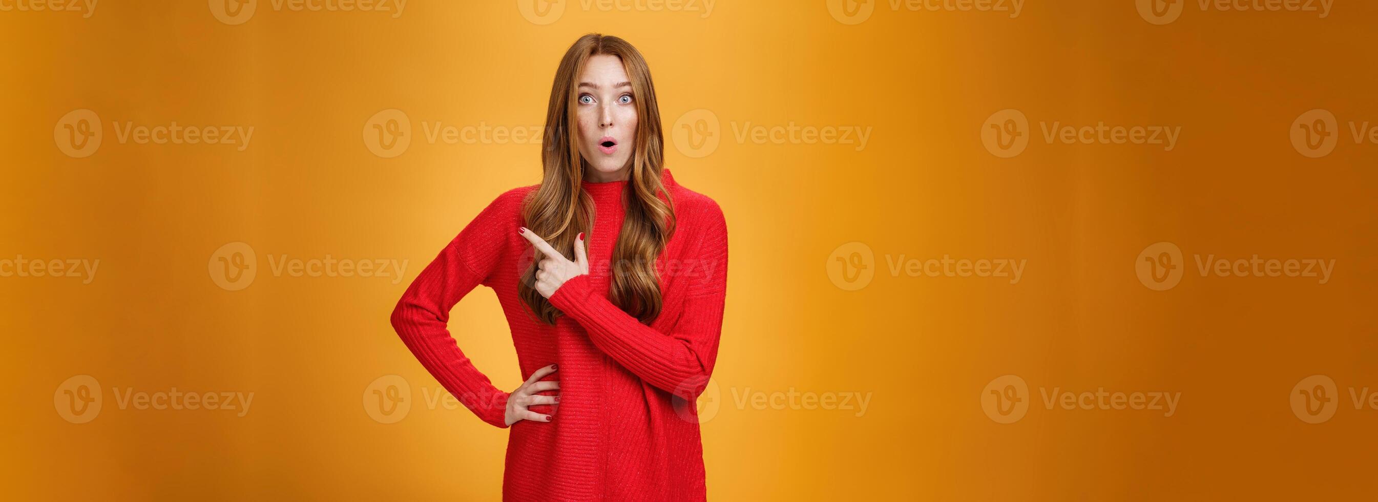 Impressed and surprised european ginger girl in red warm sweater open mouth from excitement and amazement pointing at upper right corner, gasping astonished and curious, asking question photo