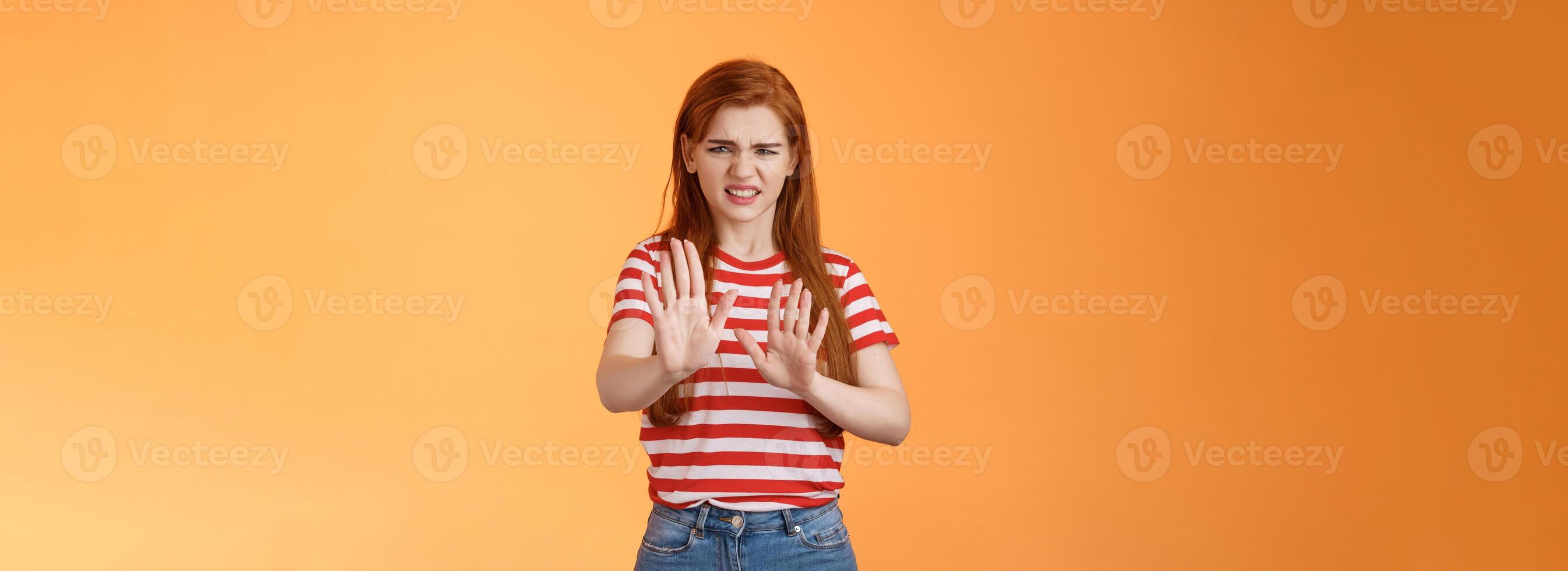 Gosh it stinks. Disgusted redhead picky woman blocking sign raise hands up defensive, grimacing, cringe from aversion awful smell, show refusal rejecting disgusting offer, stand orange background photo